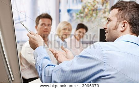 Picture or Photo of Young businessman speaking on business presentation in meeting room, pointing with pen, explaining, side view.