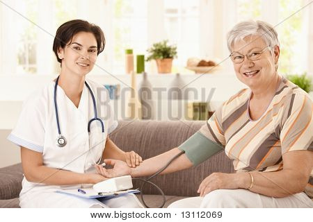 Picture or Photo of Nurse measuring blood pressure of senior woman at home. Looking at camera, smiling.?