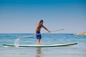 picture of oared  - The beautiful brawny guy with an oar in hands on a surfboard - JPG 